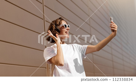 Happy young woman in stylish sunglasses taking selfie photo on smartphone on sunny summer day. Female blogger uses mobile phone camera, attractive caucasian girl touching hair for selfie picture. 112338834