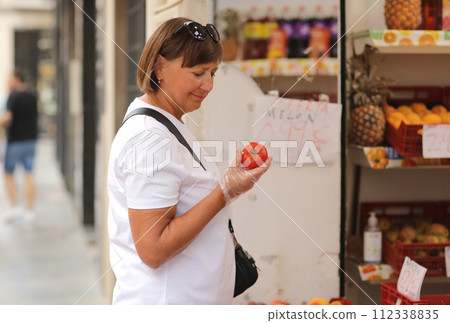 overweight woman in disposable plastic gloves chooses fresh red tomatoes, buys vegetables in a grocery store or supermarket. Concept of shopping groceries, healthy lifestyle, hygiene. 112338835