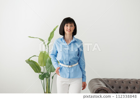 Women equality day. Smiling successful businesswoman in elegant blue shirt and white pants looks at camera at modern comfortable light office with green plant and sofa. Work life balance concept. Women equality day. Smiling successful businesswoman in elegant blue shirt and white pants looks at camera at modern comfortable light office with green plant and sofa. Work life balance concept. 112338848