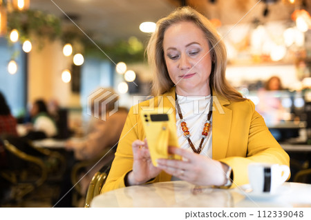Woman sits at table in restaurant, drinks coffee and writes a message on phone. 112339048