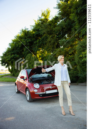 Businesswoman raises her finger while standing on country road needs help with her broken car Businesswoman raises her finger while standing on country road needs help with her broken car 112339611