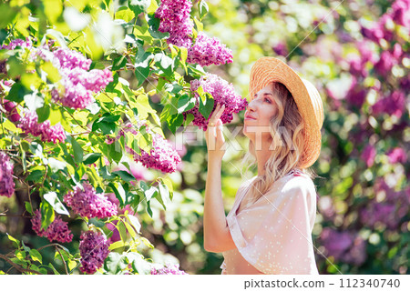 Young Woman In Summer Hat Enjoying The Fragrance Of Lilac Blooms On Sunny Day. lady in a straw hat and dress smelling lilac flowers in a sunlit garden. Young Woman In Summer Hat Enjoying The Fragrance Of Lilac Blooms On Sunny Day. lady in a straw hat and dress smelling lilac flowers in a sunlit garden. 112340740