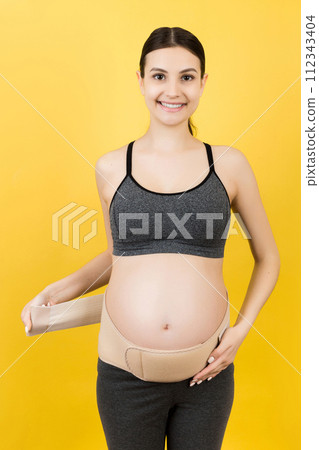 Portrait of pregnant woman dressing pregnancy corset on the third trimester at yellow background with copy space. Orthopedic abdominal support belt concept Portrait of pregnant woman dressing pregnancy corset on the third trimester at yellow background with copy space. Orthopedic abdominal support belt concept 112343404