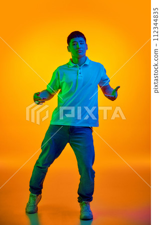 Confident young man in casual attire gesturing with open hands in vibrant neon light against gradient orange background. Confident young man in casual attire gesturing with open hands in vibrant neon light against gradient orange background. 112344835