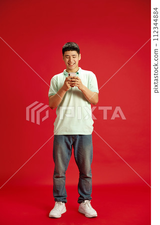 Full-length portrait of Asian man dressed casual outfit and smiling while messaging against red studio background. 112344884