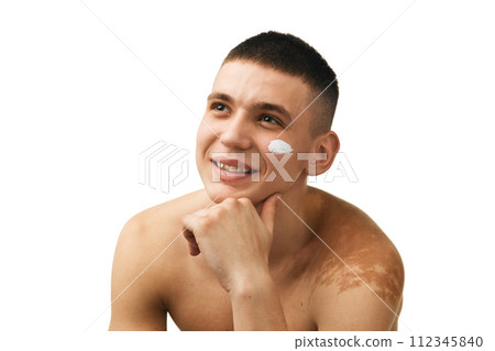 Smiling, handsome young man standing shirtless, applying face moisturizing cream isolated over white studio background 112345840