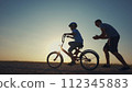 family play in the park. father teaching son to ride a bike. happy family kid dream concept. son learn to ride a bike silhouette. father supporting sunset child son riding bike summer in the park 112345883