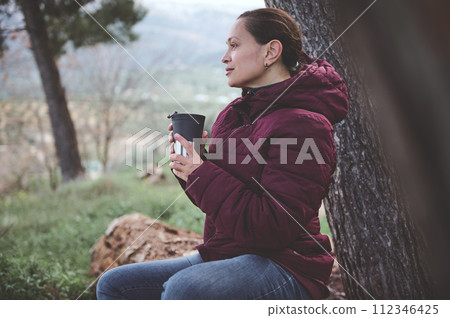 Young woman chilling out in the forest nature, holding cup of hot drink and dreamily looking into the distance Young woman chilling out in the forest nature, holding cup of hot drink and dreamily looking into the distance 112346425