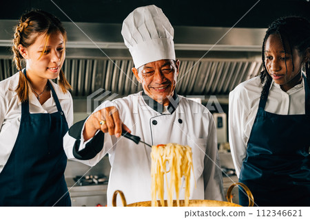 In kitchen chef teaches students. Schoolgirls in uniform make ramen soup. Kids at stove with teacher. Smiling portrait of learning is modern education. Making dinner with ladle is joyful. In kitchen chef teaches students. Schoolgirls in uniform make ramen soup. Kids at stove with teacher. Smiling portrait of learning is modern education. Making dinner with ladle is joyful. 112346621