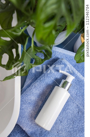 One pump cosmetic bottle on blue towels near basin and green monstera in bathroom, mockup 112346784