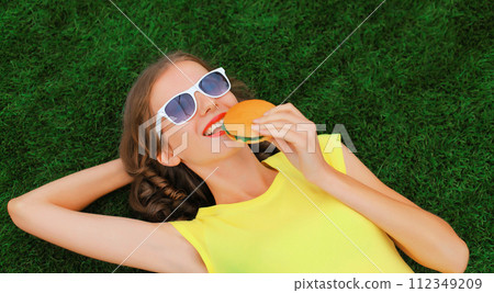 Portrait of happy smiling young woman with burger fast food lying on the grass in summer park Portrait of happy smiling young woman with burger fast food lying on the grass in summer park 112349209