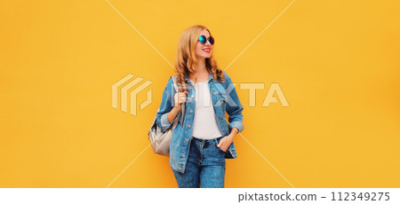 Portrait of stylish modern smiling young woman with backpack in denim jacket on yellow background 112349275