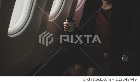 Blonde female tourist checking incoming notification on smartphone sitting on seat of airplane with netbook.Young businesswoman share media from telephone on laptop computer during plane flight Blonde female tourist checking incoming notification on smartphone sitting on seat of airplane with netbook.Young businesswoman share media from telephone on laptop computer during plane flight 112349449