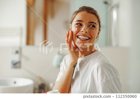 Young beautiful woman taking care of skin by applying moisturizer cream in bathroom 112350060