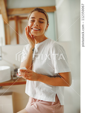Happy woman applying hydrating moisturizer on her face sitting in bathroom. Home beauty routine 112350067