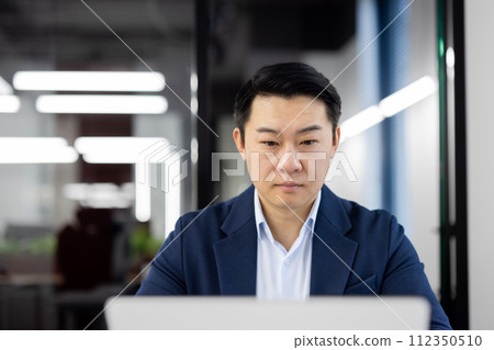 Serious businessman working diligently at his laptop in a modern office setting, displaying focus and determination. Serious businessman working diligently at his laptop in a modern office setting, displaying focus and determination. 112350510
