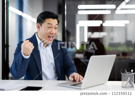 Excited male entrepreneur expressing joy for his success in a corporate setting, with a laptop in a bright office. 112350512