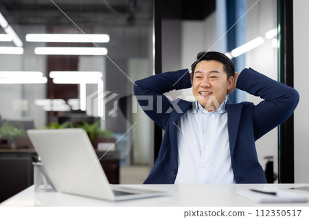 Smiling Asian businessman in a modern office leans back in satisfaction after accomplishing his work goals. 112350517