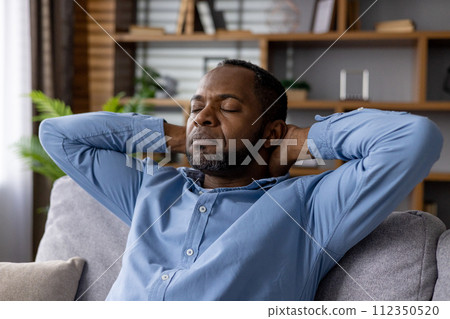 Serene African American man with closed eyes, enjoying tranquility while sitting on a sofa in a cozy living room setting. 112350520