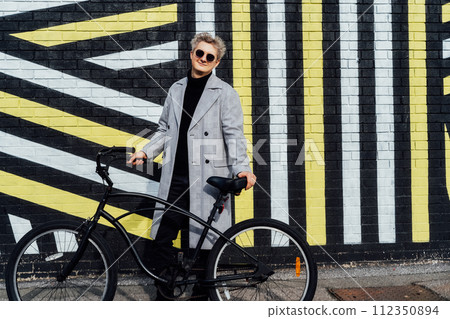 Stylish male in checked coat and sunglasses standing with retro bicycle by painted urban wall in sunny day. Neutral carbon footprint transportation. Green eco friendly mobility sustainable transport 112350894