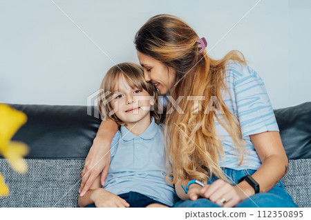 Portrait of happy smiling family at home. Son and mother sitting on the sofa, hugging each other, mom kissing son during home family weekend. Selective focus. 112350895