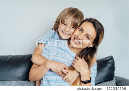 Portrait of happy smiling family at home. Son and mother sitting on the sofa and looking at camera, hugging each other during home family weekend. Selective focus 112350896