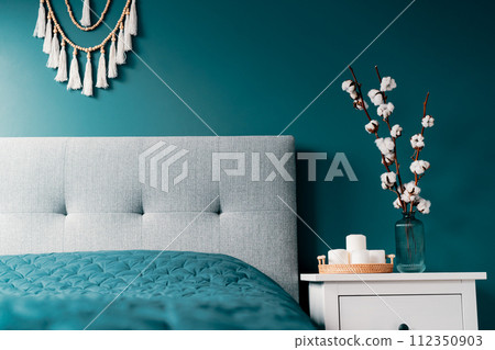 Stylish modern bedroom in dark colors. Cozy interior with blue turquoise walls, home decor. Bed with gray fabric headboard, blue blanket, bedside table, vase with natural cotton flowers, candles. 112350903