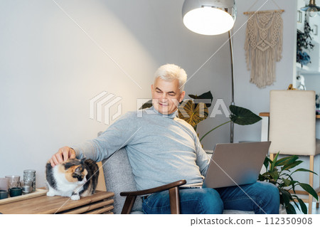 Relaxed gray hair middle-aged man stroking cat pet while working on laptop sitting in armchair at home. Cozy workspace in modern interior. Remote work at home. Senior entrepreneur manage business 112350908