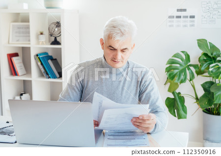 Middle aged businessman reading paper documents and using laptop while working at home office. Confident, experienced senior male professional. Small business entrepreneur manage business paperwork 112350916