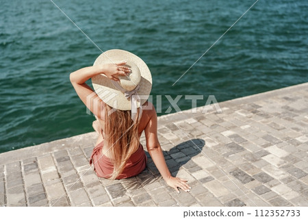 A woman in a swimsuit sits with her back holding a hat, looks at the ocean, sunny day, relaxes. 112352733