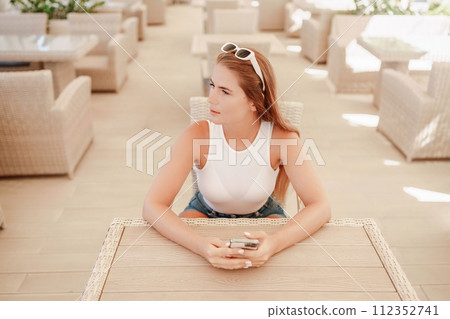Woman sits in outdoor cafe at wooden table, holds smartphone. Bright sunny day provides natural lighting. Cafe offers relaxed atmosphere for customers to enjoy refreshments. Woman sits in outdoor cafe at wooden table, holds smartphone. Bright sunny day provides natural lighting. Cafe offers relaxed atmosphere for customers to enjoy refreshments. 112352741