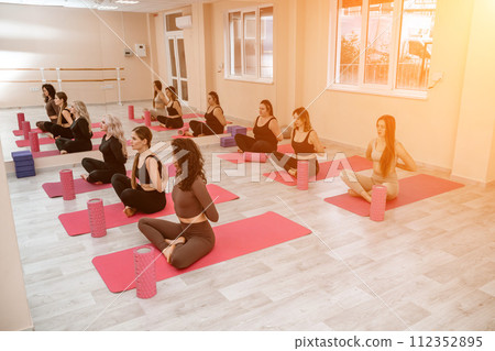 A group of six athletic women doing pilates or yoga on pink mats in front of a window in a beige loft studio interior. Teamwork, good mood and healthy lifestyle concept. A group of six athletic women doing pilates or yoga on pink mats in front of a window in a beige loft studio interior. Teamwork, good mood and healthy lifestyle concept. 112352895
