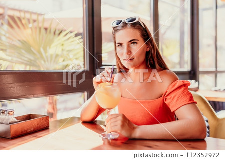 woman in a cafe seated, holding beverage. Dressed in an orange dress with her hair down. Bright interior, large windows allow natural light. Beverage consumption for refreshment. woman in a cafe seated, holding beverage. Dressed in an orange dress with her hair down. Bright interior, large windows allow natural light. Beverage consumption for refreshment. 112352972