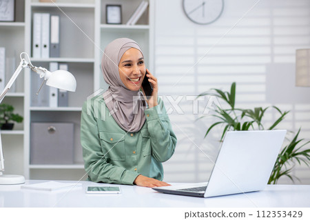 Smiling young Muslim woman wearing hijab engaged in a pleasant conversation on her smartphone at work, with laptop open. Smiling young Muslim woman wearing hijab engaged in a pleasant conversation on her smartphone at work, with laptop open. 112353429