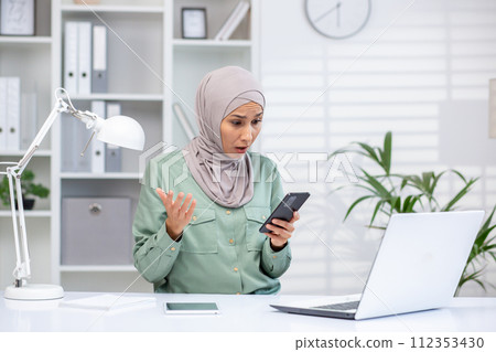 Muslim professional in hijab with shocked expression looking at smartphone, laptop and office supplies on desk. Muslim professional in hijab with shocked expression looking at smartphone, laptop and office supplies on desk. 112353430
