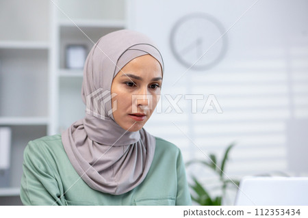 Focused Muslim woman wearing a hijab experiencing concern or stress while working in a modern office setting. Focused Muslim woman wearing a hijab experiencing concern or stress while working in a modern office setting. 112353434