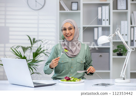 A joyful Muslim woman wearing a hijab eats a fresh salad at her desk, taking a break from work with her laptop and lamp beside. 112353442