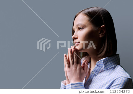 Close-up profile view of young woman with folded hands on gray background, copy space 112354273
