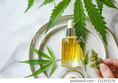 Hand holding dried cannabis bud, cosmetic oil in dropper bottle on plate with hemp bud and leaves, light marble background. Aesthetic background with medical marijuana in beauty industry CBD, CBN oil 112355276