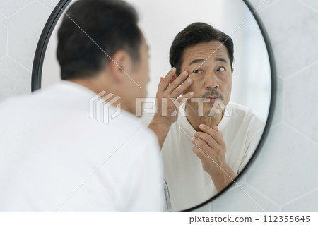A man in his mid-50s covers the corners of his eyes while looking in the mirror A man in his mid-50s covers the corners of his eyes while looking in the mirror 112355645