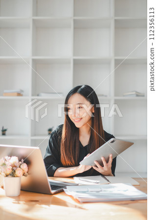 A smiling professional woman utilizes a digital tablet alongside her laptop, embracing technology in a modern, light-filled workspace, indicative of a dynamic and connected work style. A smiling professional woman utilizes a digital tablet alongside her laptop, embracing technology in a modern, light-filled workspace, indicative of a dynamic and connected work style. 112356113