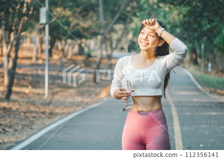 A radiant fitness enthusiast wipes sweat from her brow while holding a water bottle during a jogging break, set against a serene park backdrop, embodying active wellness. A radiant fitness enthusiast wipes sweat from her brow while holding a water bottle during a jogging break, set against a serene park backdrop, embodying active wellness. 112356141