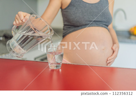 Embracing the vital benefits of water during pregnancy, a pregnant woman stands in the kitchen with a glass, highlighting hydration's crucial role in maternal well-being Embracing the vital benefits of water during pregnancy, a pregnant woman stands in the kitchen with a glass, highlighting hydration's crucial role in maternal well-being 112356312