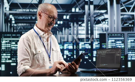 Old system administrator in data center, solving tasks on tablet while running scripts on laptop terminal. Qualified employee amidst servers, accessing databases and manipulating lines of code 112356369