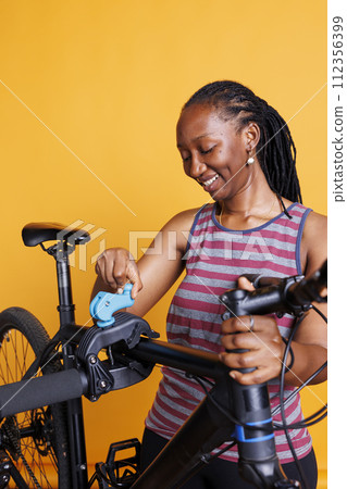 African american female cyclist inspects her broken bicycle frame on repair stand against isolated yellow backdrop. Black woman preparing to make adjustments and repairs with specialized equipment. 112356399
