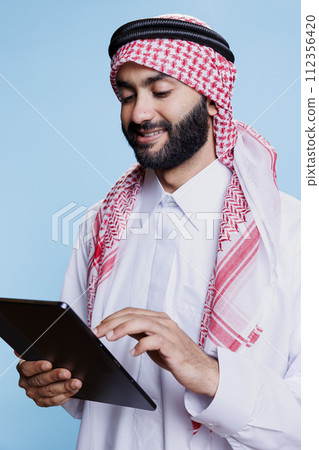 Smiling muslim person dressed in traditional clothes using digital tablet while engaging with social media messages. Cheerful arab person in islamic thobe and headdress using portable device in studio 112356420