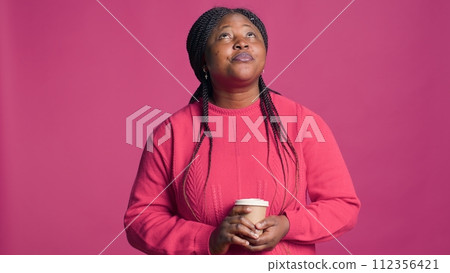 Portrait of african american woman in thoughts looking up and holding cup of coffee. Female fashion blogger in contentment wearing pink sweater enjoys hot beverage. 112356421