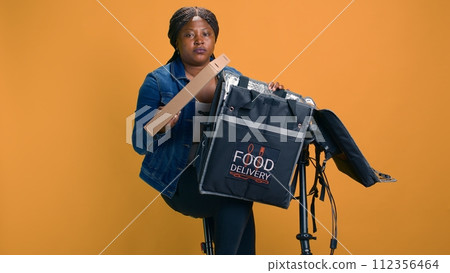 Black woman on bicycle carefully delivering fresh takeout meal with courier bag. African american delivery person providing professional customer service by making efficient delivery. 112356464