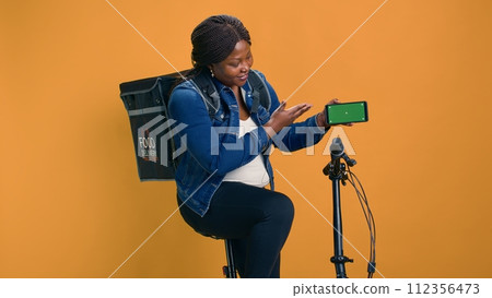 African american courier gripping cell phone showing green screen display while riding bicycle. Black woman demonstrates cutting-edge delivery app on mobile device with isolated mockup template. 112356473