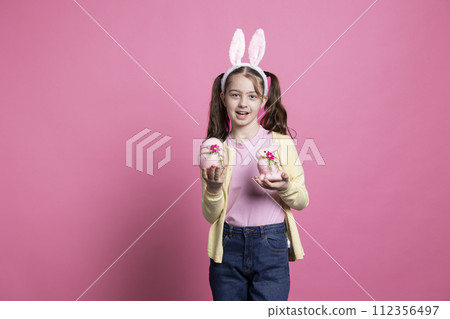 Small girl with bunny ears holding pink easter decorations in front of camera, presenting her handmade egg and stuffed rabbit. Young child smiling in studio and celebrating spring festivity. Small girl with bunny ears holding pink easter decorations in front of camera, presenting her handmade egg and stuffed rabbit. Young child smiling in studio and celebrating spring festivity. 112356497